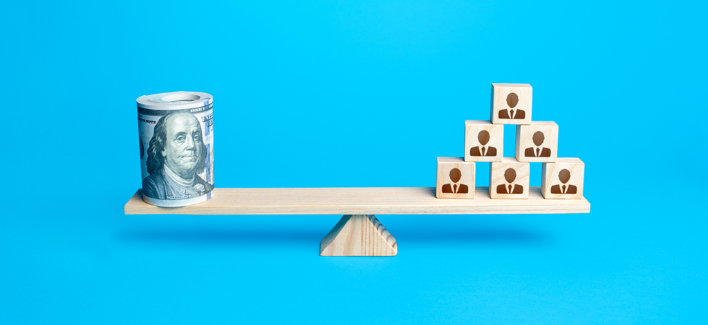 Wooden balance block with roll of bills on one side and silhouettes of people on the other