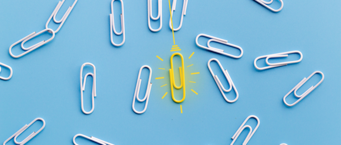 One yellow paperclip standing out among a group of white paperclips on a light blue background